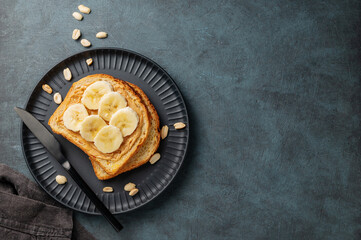 Peanut butter toast and banana on a black plate on a dark background with nuts, knife and napkin.