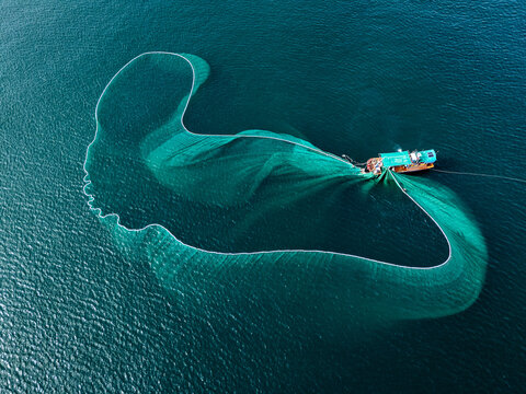 Aerial view of a fishing boat amidst the vast, deep blue sea, casting a wide net like a shimmering emerald veil across the water, An Ninh Dong, Phu Yen, Vietnam.