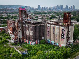 Aerial view of weathered grain elevators adorned with vibrant graffiti, rising amidst lush greenery against the backdrop of the Montreal skyline, Montreal, Quebec, Canada.