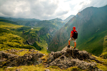 A man with a backpack wearing a red T-shirt and a cap is standing on a cliff in the Caucasus, looking into the distance.
