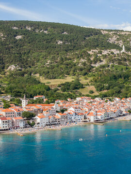Aerial view of the sun-kissed shore where the turquoise sea meets the red-roofed houses nestled against the verdant hills, Baska, Primorje-Gorski Kotar County, Croatia.