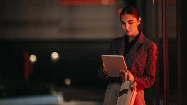 A poised professional woman stands outdoors at night, examining content on a tablet. The red ambient lighting adds a modern, urban business vibe.