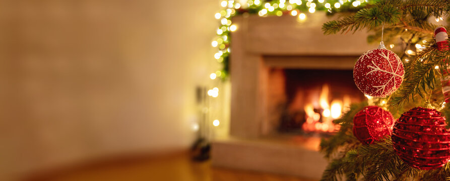 Festive red ornaments hang upon the Christmas tree beside a glowing fireplace, creating a cozy holiday mood