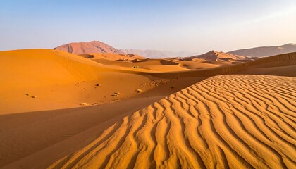 Rippled sand dunes under a bright sky, leading toward distant mountains under clear sky, a wide panorama