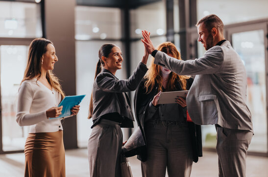 Diverse group of people in business attire celebrate, sharing a high five in a bright lobby. They stand with tablets and documents, conveying teamwork, collaboration, and positive corporate culture.