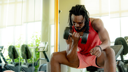 A determined man with dreadlocks in a red tank top intensely performing bicep curls with a heavy dumbbell during his gym workout. Bodybuilding concentration fitness journey and physical activity, heal