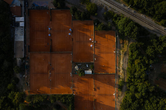 Aerial view of ochre tennis courts nestle against verdant trees, casting stark shadows under the sun's gaze, Rijeka, Primorje-Gorski Kotar County, Croatia.