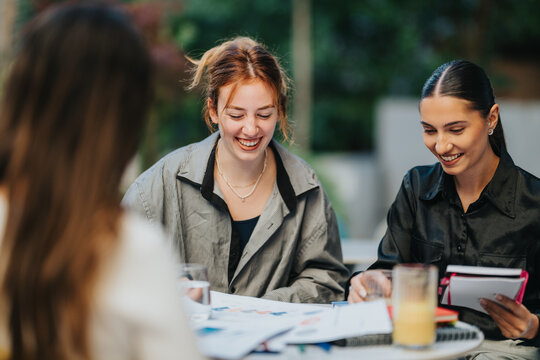 Three people sit at a round table outside, smiling as they review papers and notes. A casual business meeting among friends and colleagues, with drinks and a notebook in a relaxed setting. - Powered by Adobe