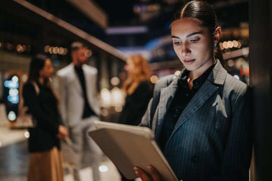 A focused woman in a pinstripe blazer reviews information on a tablet in a contemporary office setting. Blurred coworkers in the background suggest collaboration and networking during a business day.