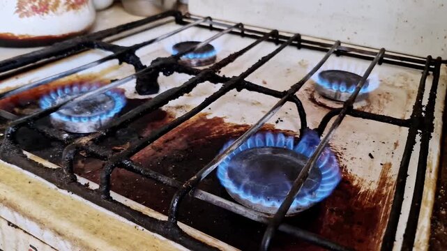 Overview of multiple blue flames burning on an old, dirty gas stove. Appliance has greasy grates, stained white enamel, and food residue, representing inefficient energy use in a household kitchen.