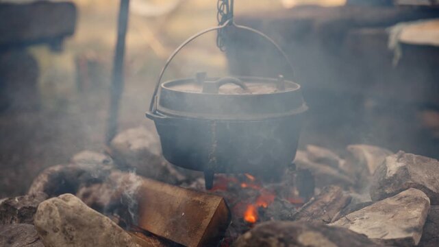 Rustic Dutch oven hanging above open campfire in forest with rising smoke and glowing wood logs