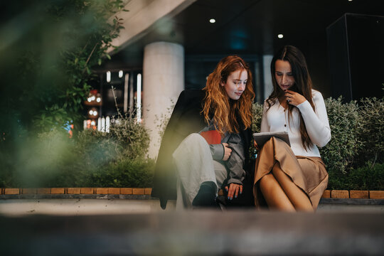 Two young women sit on a bench in an urban night scene, reviewing a tablet together. They project focus, collaboration, and entrepreneurial vibes in a stylish, professional moment.