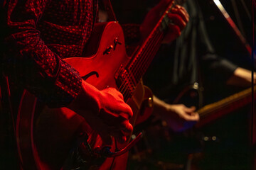 Musicians playing electric guitars under red stage lighting during a live performance. The close-up view focuses on the hands, instruments, and illuminated details of the scene.