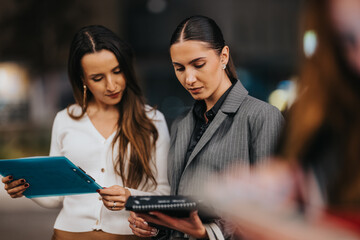 Two professional women examine folders and notebooks while standing outside, sharing ideas and...