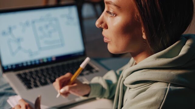 Writing notes on laptop screen. Student sketches design on notebook with pencil. Woman studies layout while laptop displays wireframe. Image shows focused learning and practical education at desk.