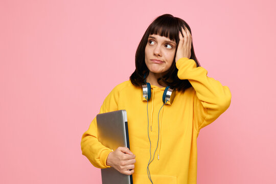 Young student in a yellow sweater holds a laptop beside a pink backdrop, headphones resting on their neck while they ponder ideas. A casual, modern learning moment with a creative, hopeful vibe.