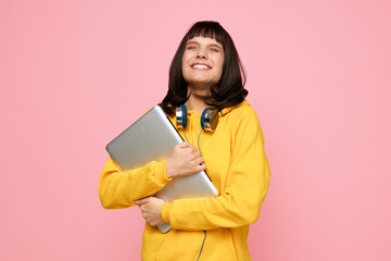 A cheerful student stands against a pink backdrop, hugging a laptop and wearing headphones. This bright, contemporary portrait conveys creativity, study, and positive energy.