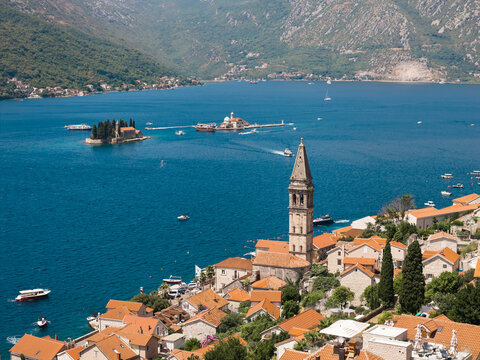 Aerial view of terracotta rooftops cascade down to the serene bay dotted with Sveti Dorde and Gospa od Skrpjela islands, framed by verdant mountains, Perast, Kotor Municipality, Montenegro.