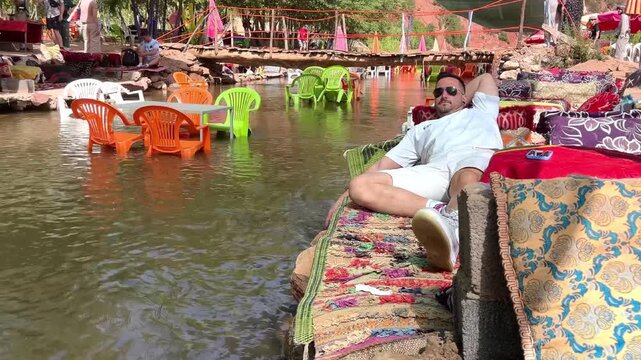 White young male tourist relaxing on a colorful Berber rug at an unconventional outdoor cafe with tables and chairs set up in the flowing river of the Ourika Valley, near Marrakech Morocco