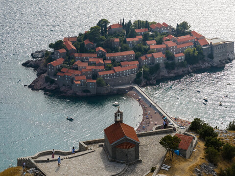 Aerial view of the terracotta rooftops of Sveti Stefan contrast with the azure Adriatic, linked by a narrow isthmus to the mainland, Sveti Stefan, Budva Municipality, Montenegro.