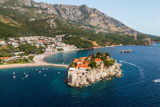 Aerial view of the terracotta rooftops of Sveti Stefan contrast against the azure Adriatic, cradled by rugged mountains and sandy beaches, Sveti Stefan, Budva Municipality, Montenegro.