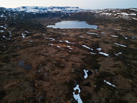 Aerial drone view of a vast mountain landscape in Norway with a remote lake, tundra, and melting snow.