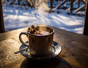 A steaming mug of hot drink with cinnamon sticks sits on a wooden table, with a snowy winter landscape visible through a window in the background.