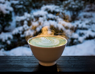 A steaming bowl of vibrant green matcha tea with intricate latte art, set against a blurred backdrop of a snowy winter landscape.