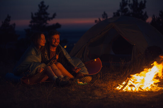 Young tourist couple exploring new places. An attractive woman and a handsome man spend time together in nature. Sitting by the campfire and tent in the evening mountains and making marshmallows.