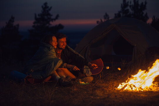 Young tourist couple exploring new places. An attractive woman and a handsome man spend time together in nature. Sitting by the campfire and tent in the evening mountains and making marshmallows.