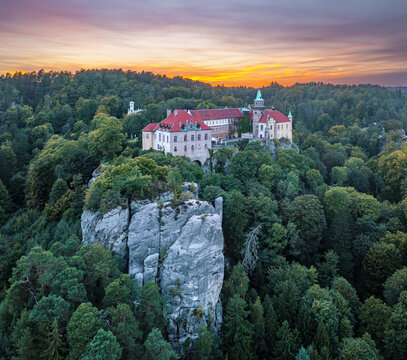 Aerial view of the castle perched atop a rocky outcrop amidst lush green forest under a vibrant sunset sky, Turnov, Liberec Region, Czechia.