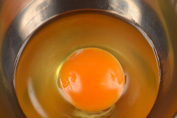 A vibrant orange raw egg yolk sits centered in a reflective stainless steel mixing bowl, capturing rich culinary color and texture
