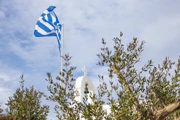 traditional Greek white chapel with bell tower, cross and fluttering flag, seen from olive tree branches. Greek aesthetic symbols. orthodox