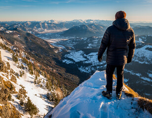 A hiker on a snow-covered summit gazes at a stunning mountain range and valley during golden hour.