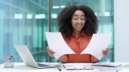 Joyful young African American businesswoman is satisfied with results of a financial report she is reviewing while sitting at a workplace in office. Female manager is happy with positive indicators - Powered by Adobe