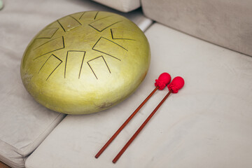 Golden handpan drum with geometric sound holes rests on a sofa, by two vibrant red mallets
