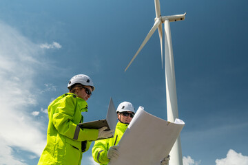 Two people are standing next to a wind turbine. One of them is holding a laptop. Low angle view of two technicians (man and woman) inspecting wind turbine structure.