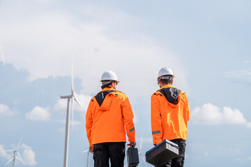 Two men in orange jackets are standing on a wind farm. One of them is holding a toolbox. Technician team in high visibility jackets carrying toolboxes for maintenance service at renewable energy site.