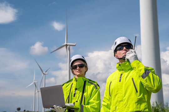 Two workers in yellow jackets and helmets are standing next to a laptop. One of them is talking on a cell phone. Low angle view of two technicians (man and woman) inspecting wind turbine structure. - Powered by Adobe