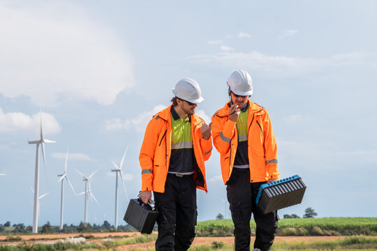 Two men in orange jackets are walking and talking. One of them is holding a toolbox. Technician team in high visibility jackets carrying toolboxes for maintenance service at renewable energy site.