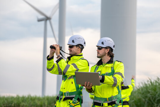 Two men in yellow jackets and hard hats are standing next to a wind turbine, one of them holding a l. Group of engineers (four people) with PPE, collaborating and walking at wind turbine field.