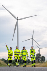 A group of four workers in yellow and black safety gear are walking near a large wind turbine. Technicians in PPE discussing blueprint and inspecting wind turbines, planning for maintenance.