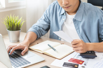 Stressed asian young businessman, employee male using laptop computer to calculate expenses, hand holding bills and receipt for to payment on table at home. Financial, finance of banking concept.