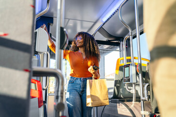 Young woman smiling, standing on a city bus and paying with a payment card for her ride. She is holding a paper bag © Koldo_Studio