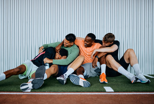 Group of fit young diverse men in sportswear laughing and messing around while sitting together on the floor of a gym after a workout class