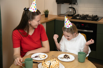 Happy mothers and daughter in party hats eating cake and drinking tea in a modern kitchen setting, sharing a special family moment