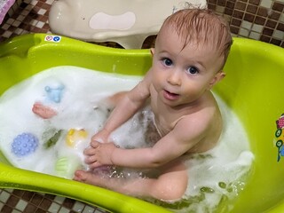 A happy seven-month-old baby sitting in a bathtub full of fluffy foam during bath time.

