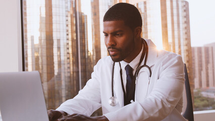 Male Doctor With Stethoscope Working On Laptop At Desk In Hospital Office