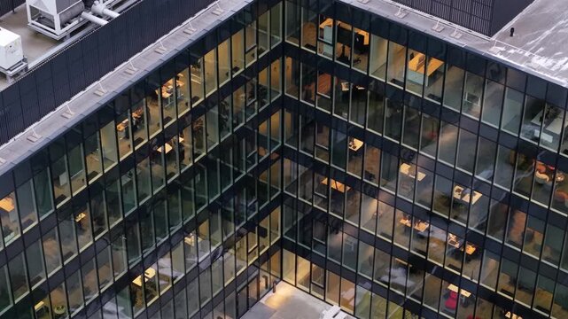 Aerial close-up of a glass-clad office building in Vilnius, Lithuania, showing illuminated workspaces and a rooftop with industrial HVAC systems and ventilation infrastructure.