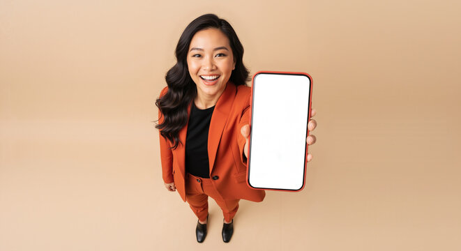 Smiling Asian woman holding a smartphone with a blank white screen for mockup, high angle view of a happy professional on a beige studio background.